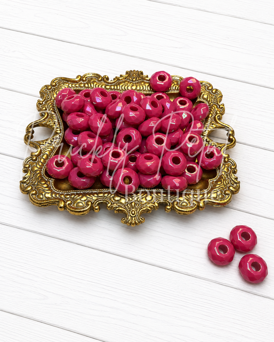 Bright pink round craft beads with center holes, displayed in an ornate gold-colored tray on a white wooden background, with three additional beads placed beside the tray.