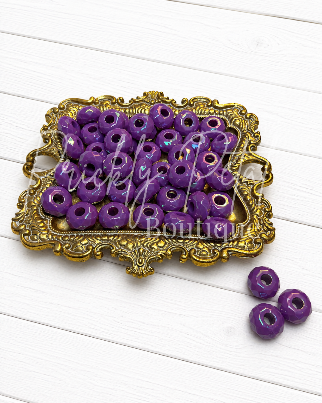 Purple round craft beads with a glossy finish and central holes, displayed on an ornate gold tray with three additional beads placed beside it on a white wood background. 