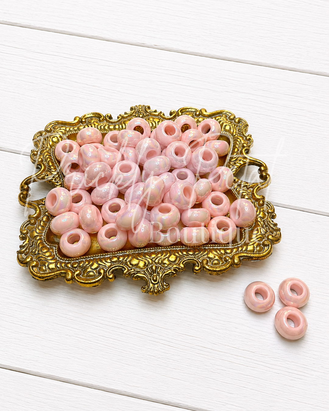 Pink round craft beads with a glossy finish, displayed on an ornate gold tray with three additional beads on the side, set against a white wooden background.