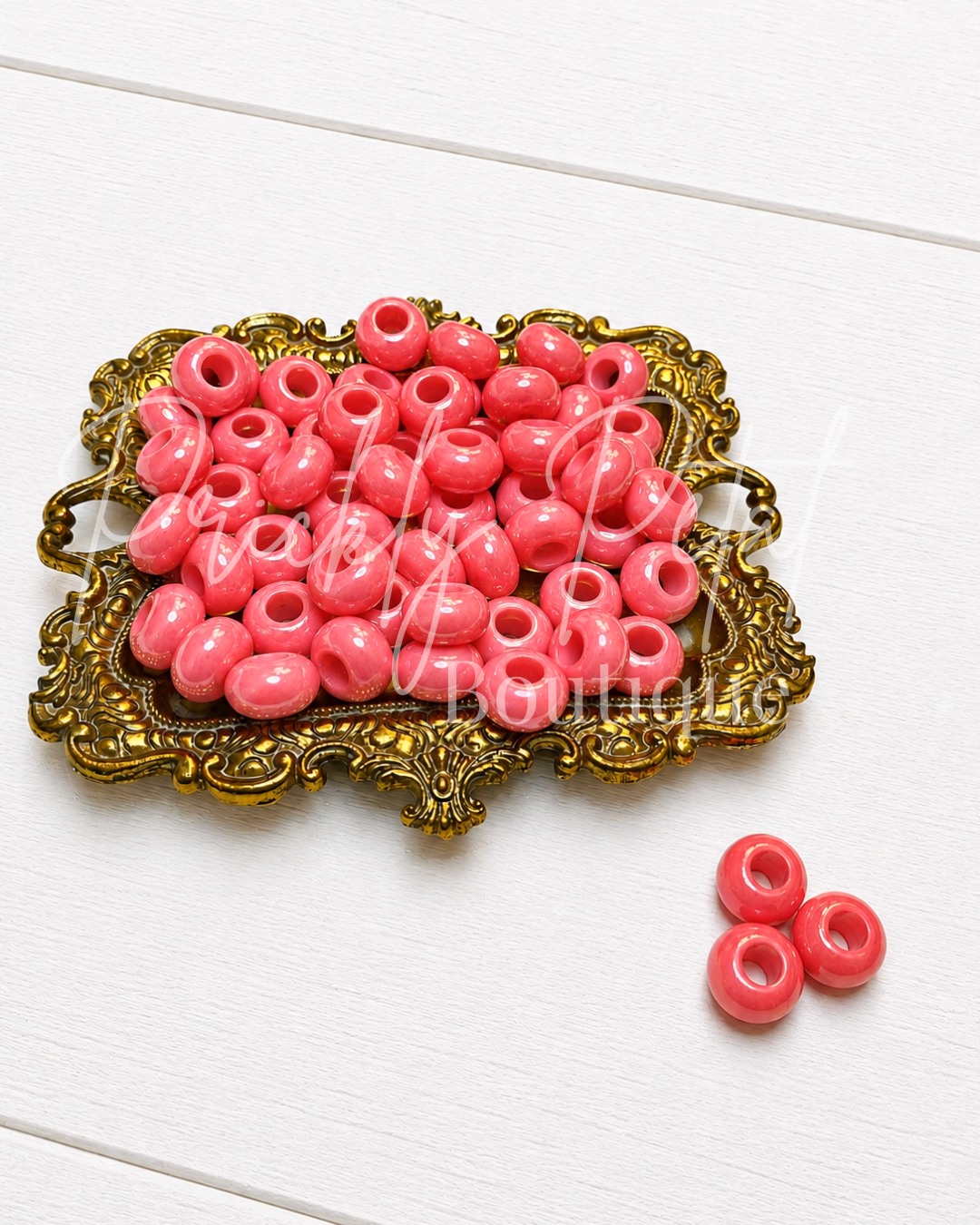 Pink round craft beads with center holes, displayed in an ornate gold tray with three additional beads on a white wood background.