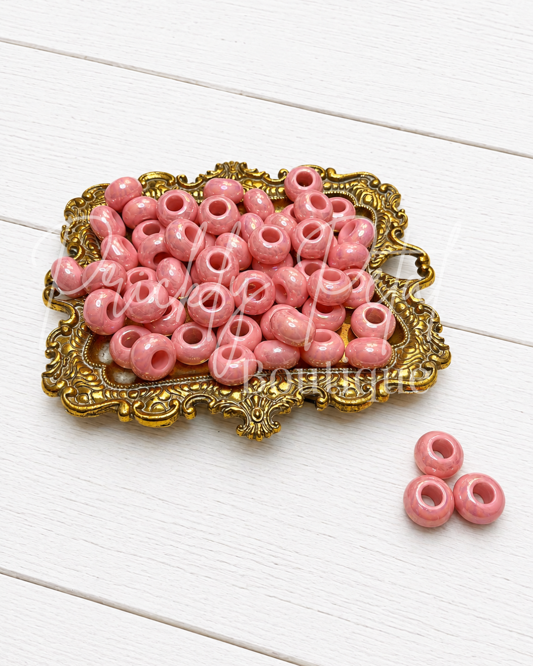 Pink wooden bead charms with a hollow center, displayed on an ornate gold tray with three additional beads on a white wooden surface.
