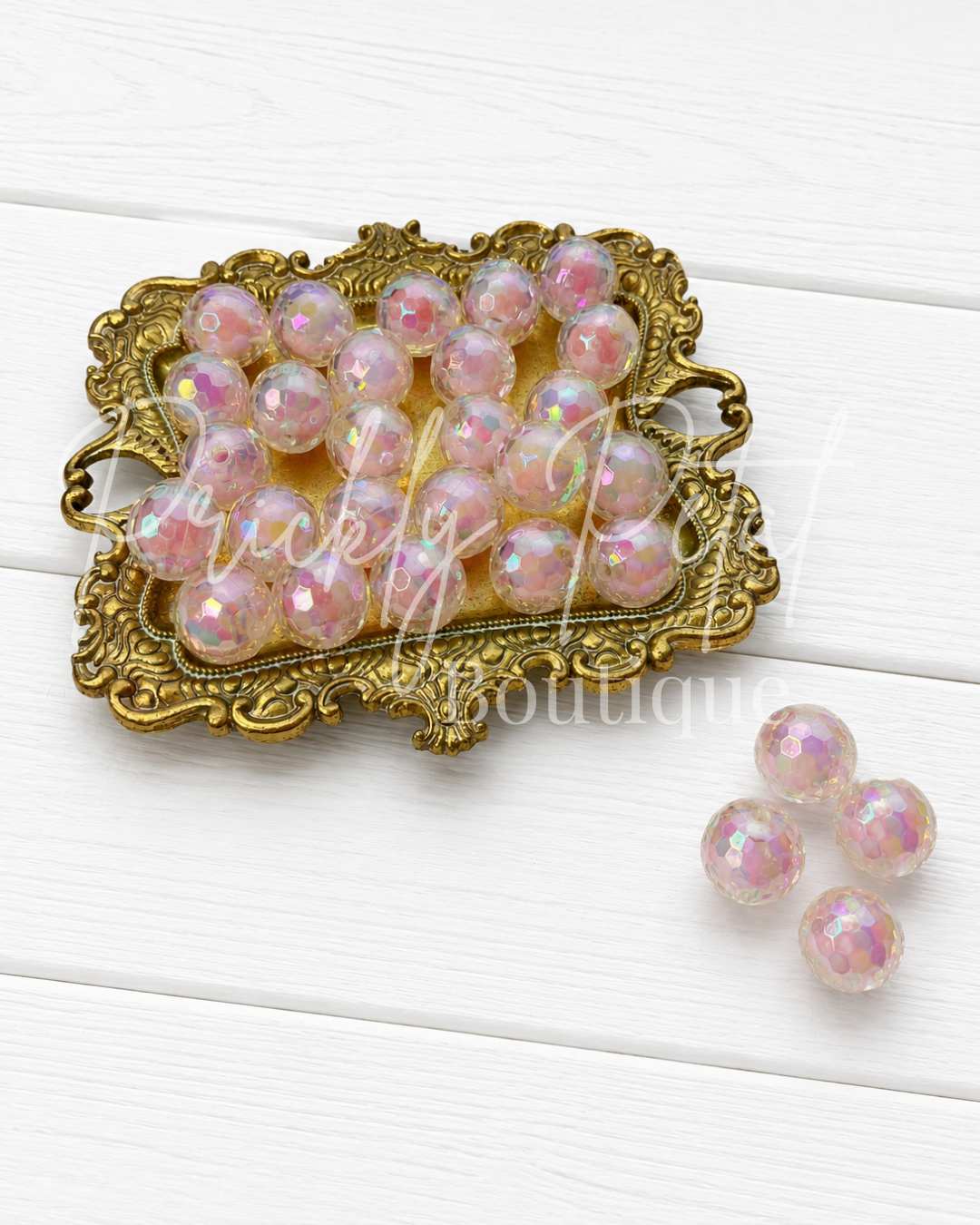 A set of iridescent purple glass beads with a faceted finish, displayed on an ornate gold tray with three additional beads placed beside it on a white wood background.