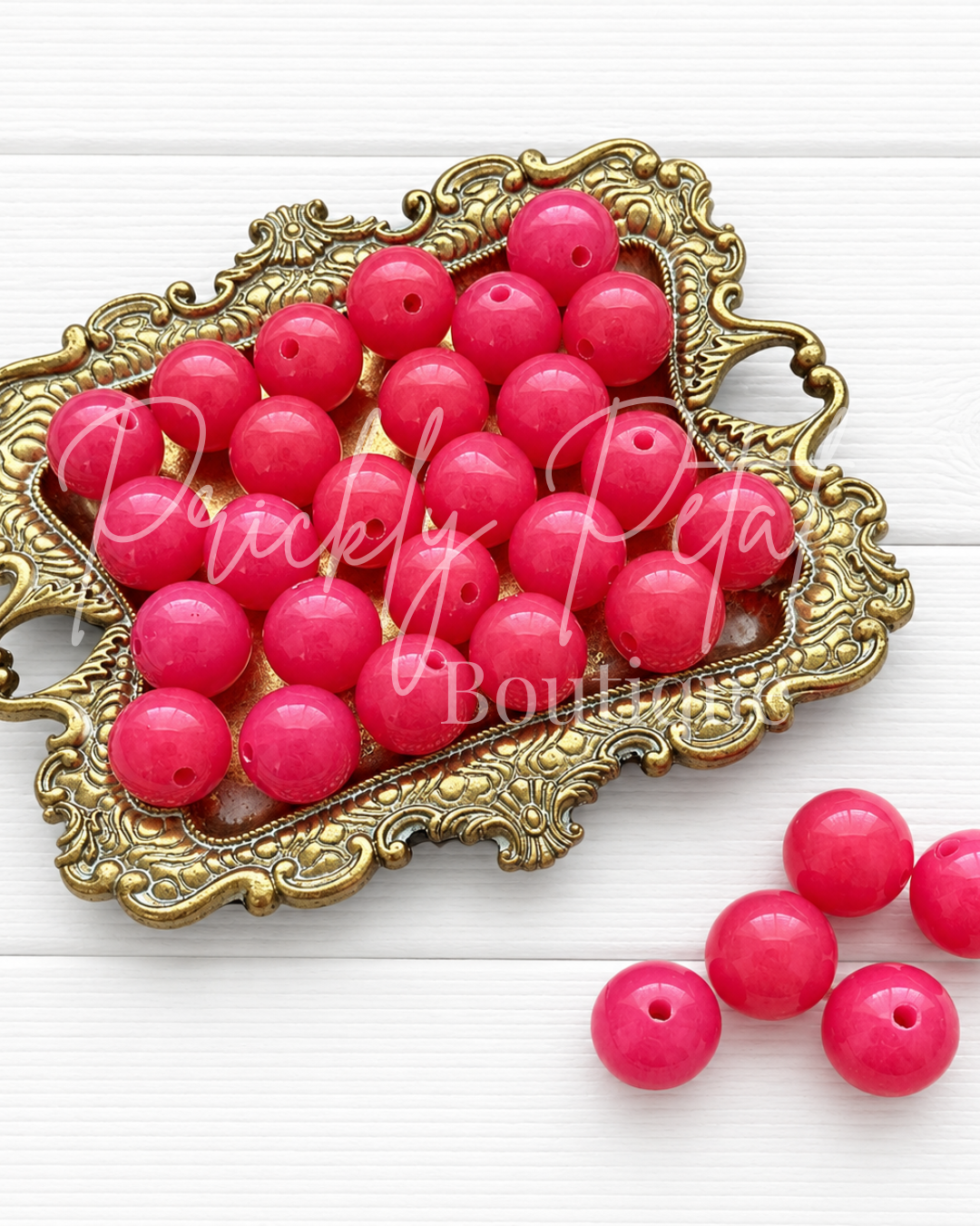 Bright pink round craft beads with holes, displayed in an ornate gold tray on a white wood background, with a few beads placed outside the tray. 
