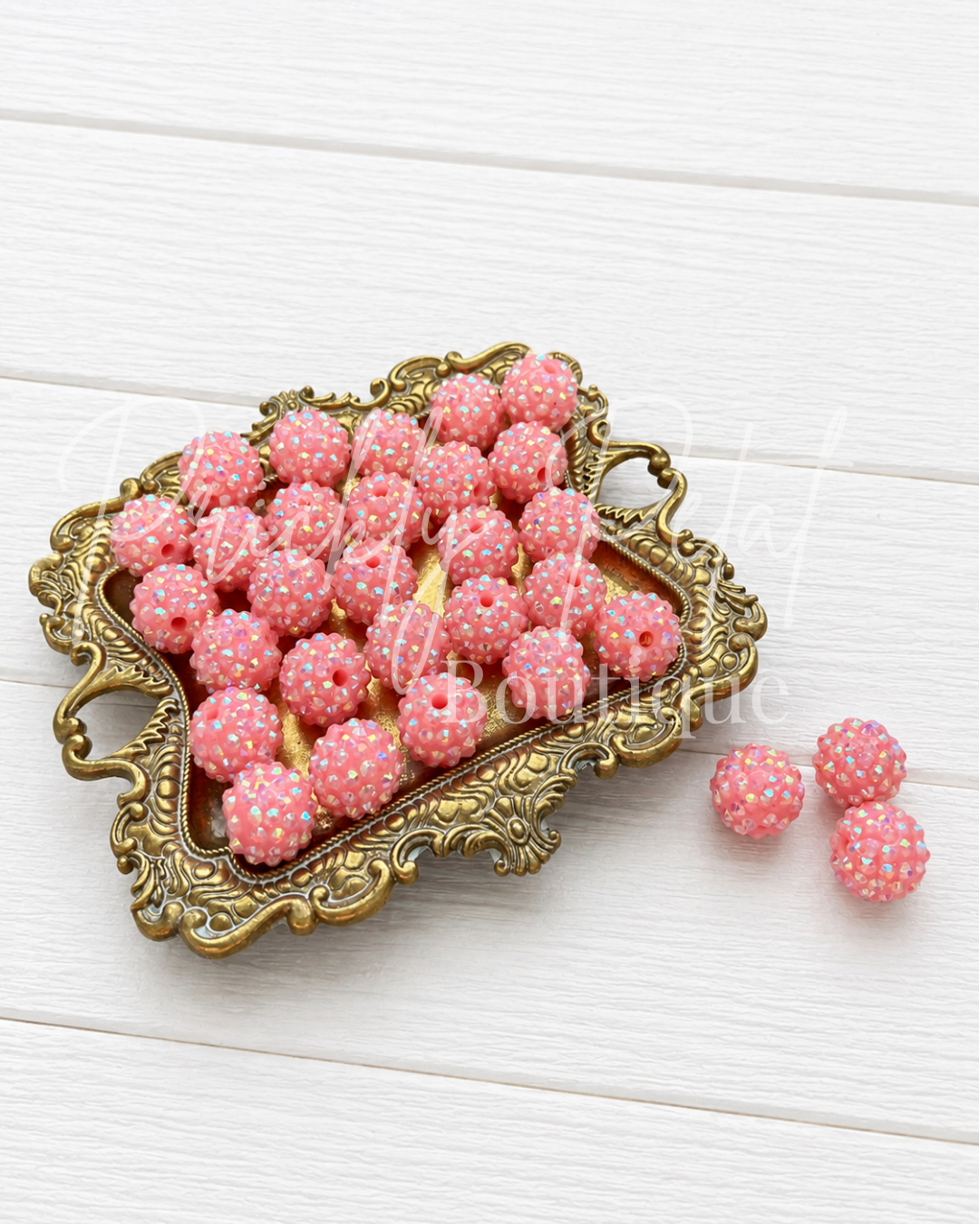 Pink and white sugar-dusted round beads arranged on an ornate gold tray, with three additional beads placed beside it on a white wood background.