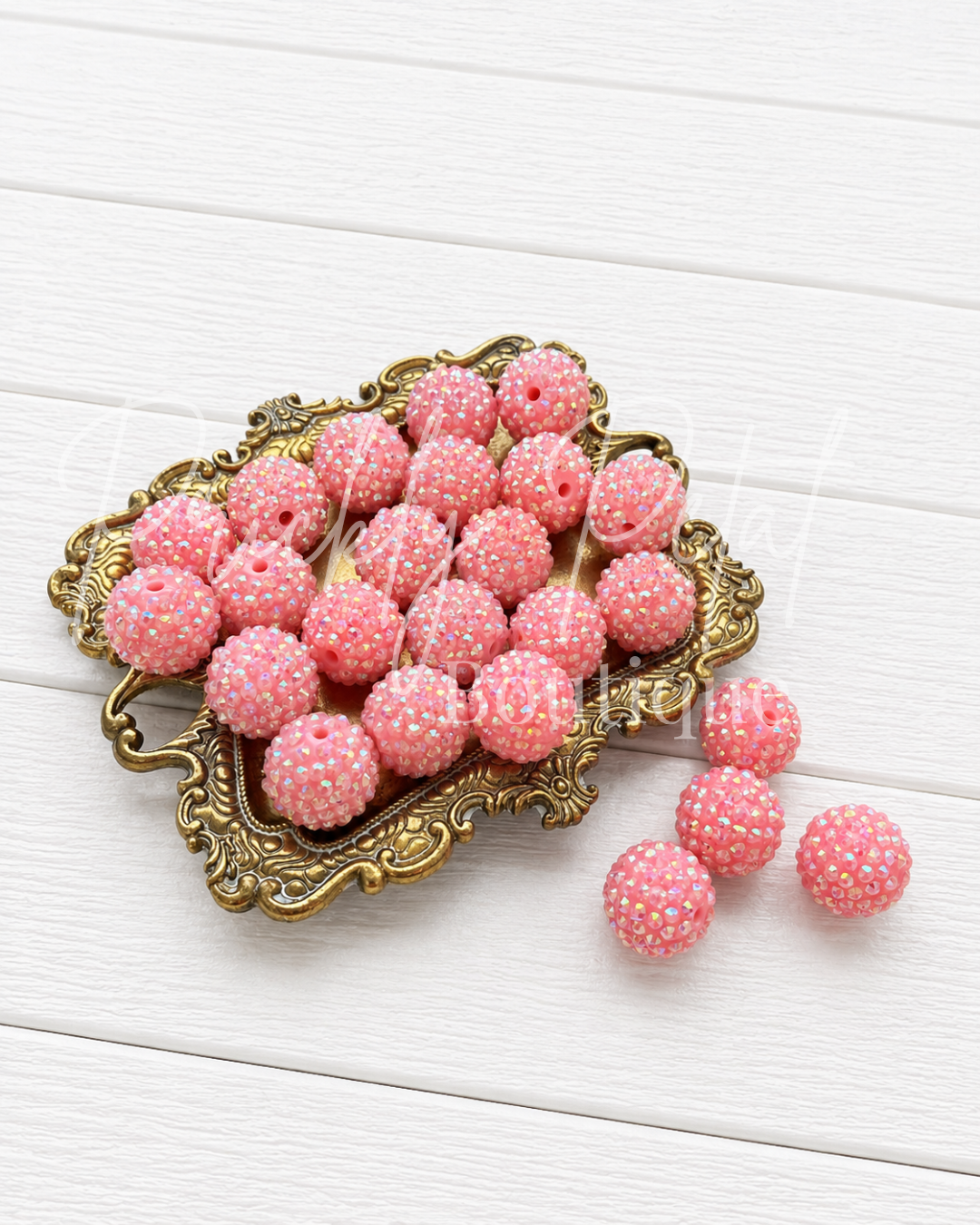 Pink sugar-dusted beads with white pearl accents, displayed on an ornate gold tray with three additional beads resting beside it on a white wooden background.