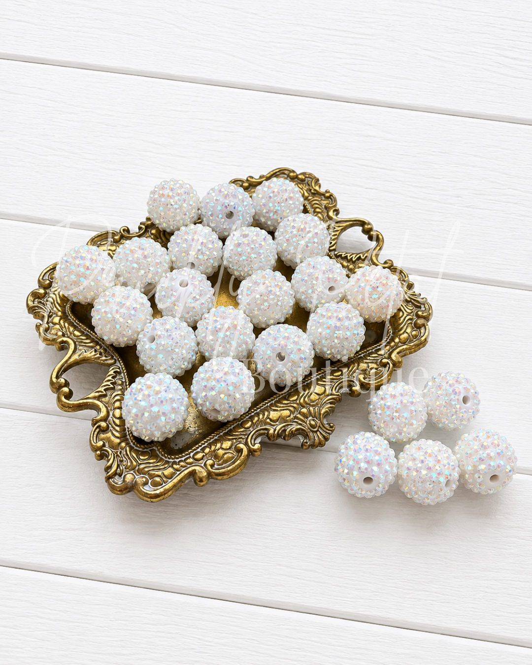 White iridescent rhinestone-covered round beads with center holes, displayed on an ornate gold tray with several beads resting beside it on a white wood plank background.