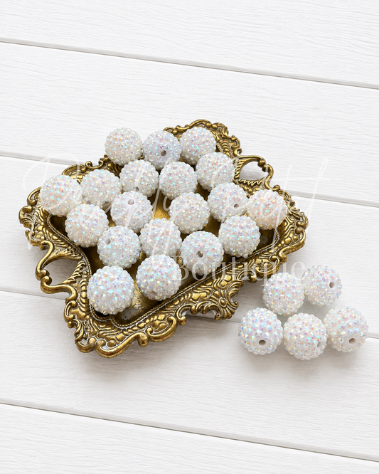 White iridescent rhinestone-covered round beads with center holes, displayed on an ornate gold tray with several beads resting beside it on a white wood plank background.