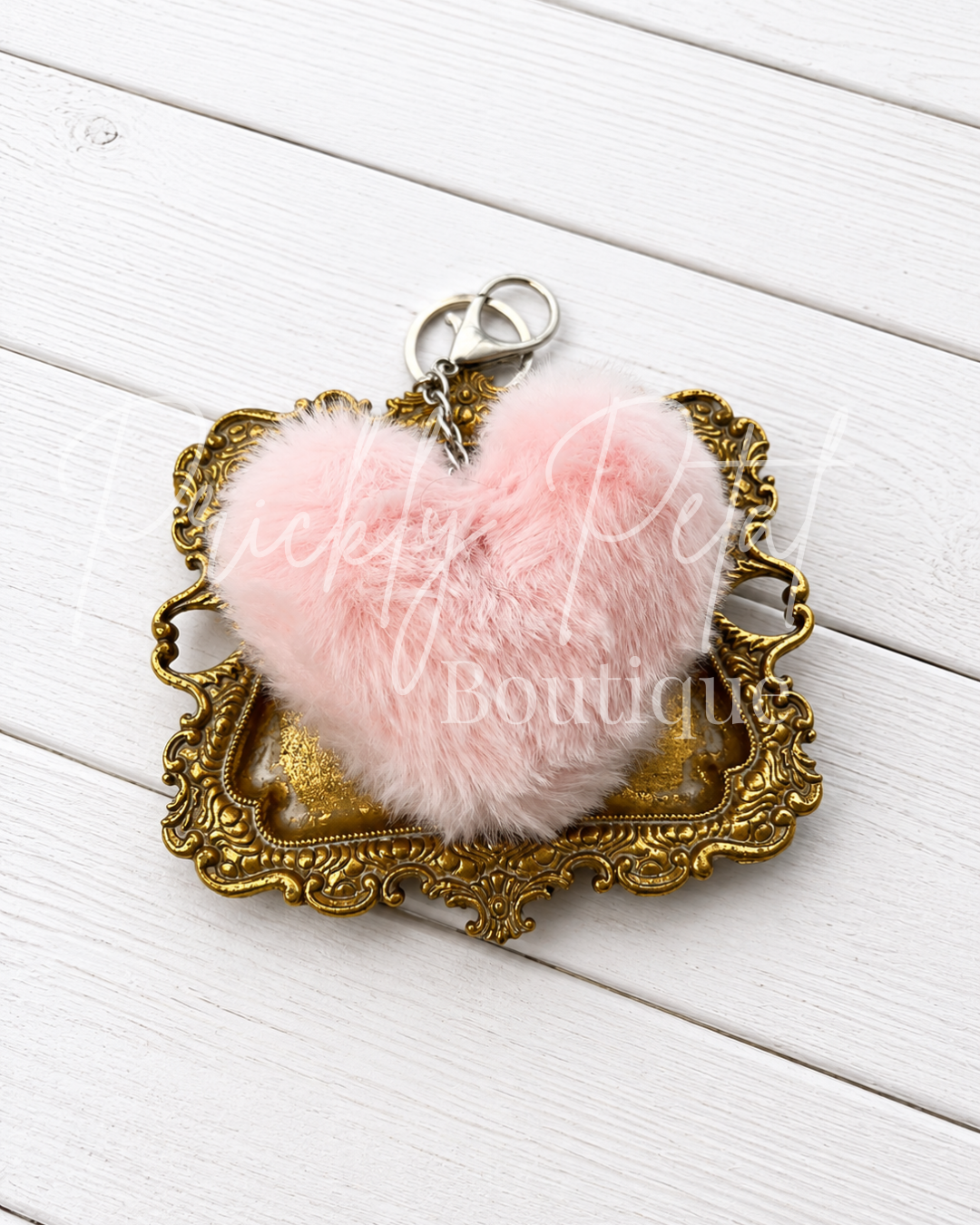 A pink fluffy heart-shaped keychain charm resting on an ornate gold-colored metal tray, set against a white wooden plank background.