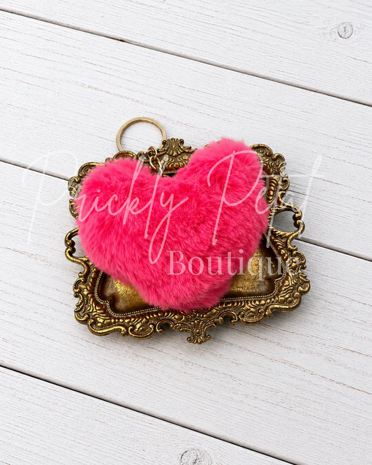 A bright pink fluffy heart-shaped keychain or bag charm resting on an ornate antique gold metal frame, set against a white painted wood plank background.