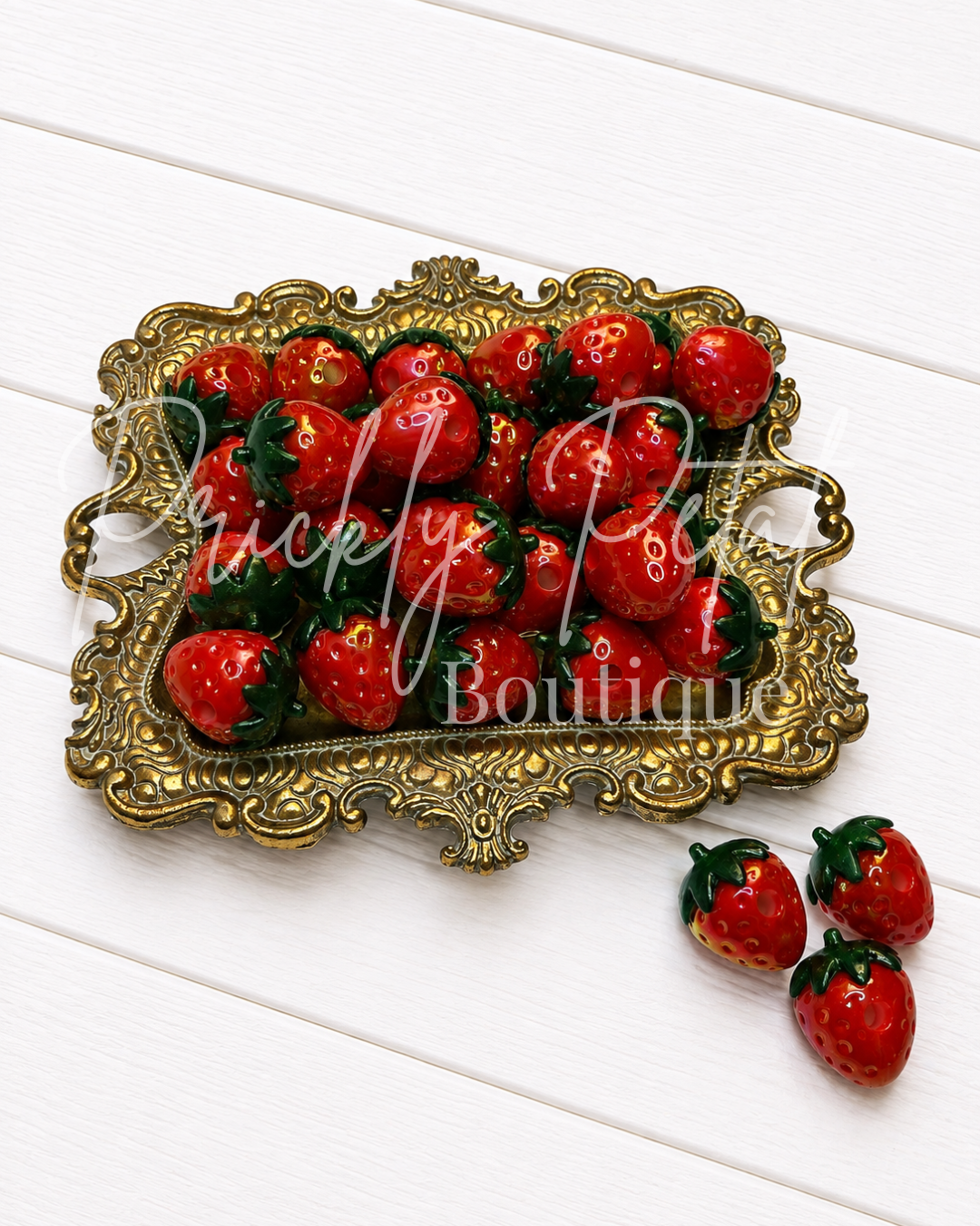 Three small red strawberries with green tops displayed on an ornate gold tray, with two additional strawberries placed beside it on a white wood plank surface. 