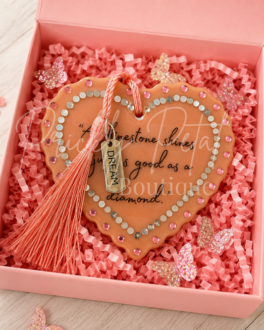 A heart-shaped peach ornament with rhinestone trim and a tassel, displayed inside a pink gift box with pink shredded paper. The ornament features cursive text and a small hanging tag that reads 'DREAM'.