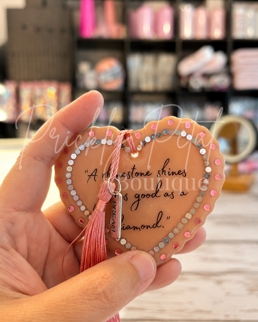 A heart-shaped peach ornament with rhinestone trim and a tassel, displayed inside a pink gift box with pink shredded paper. The ornament features cursive text and a small hanging tag that reads 'DREAM'.