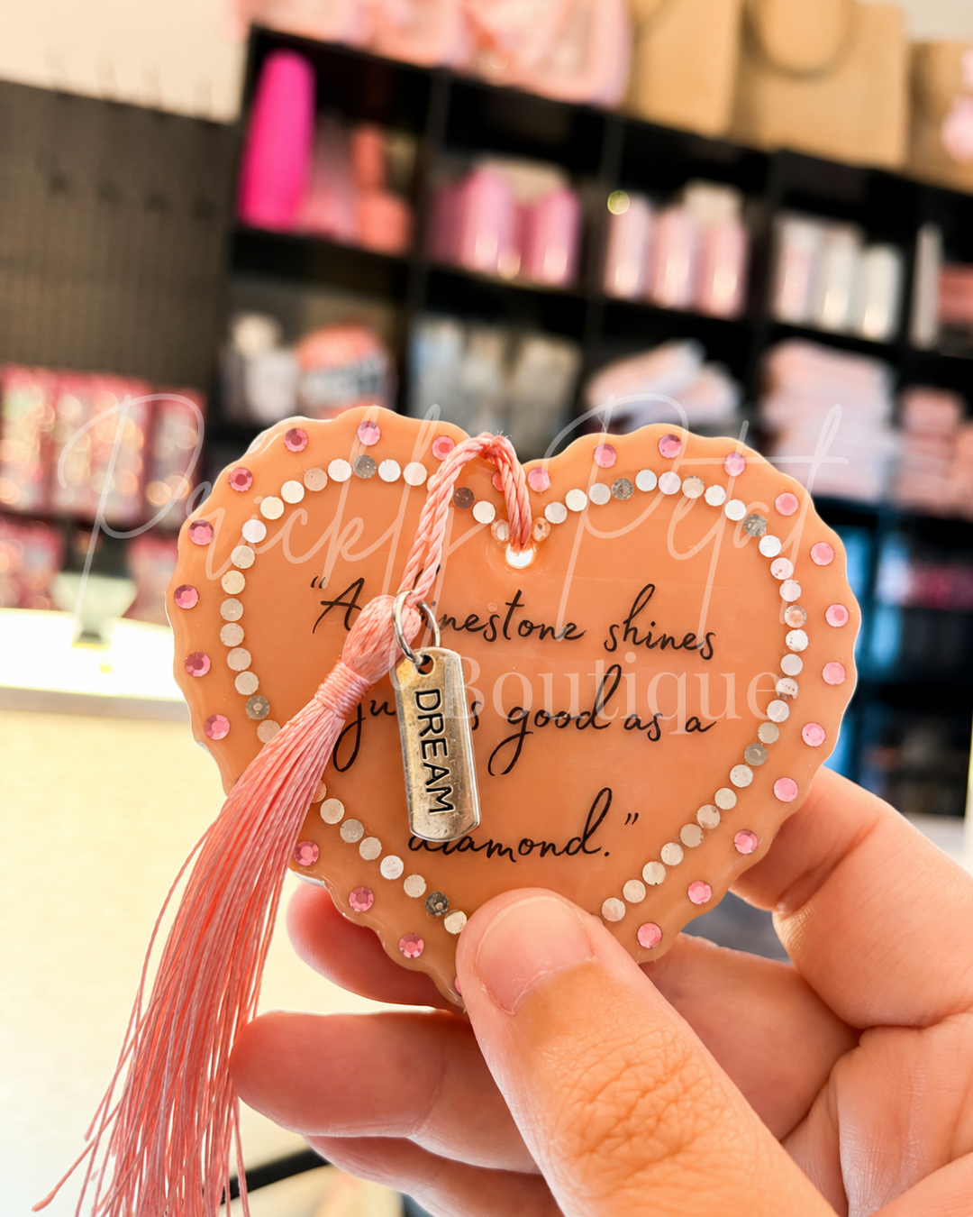 A hand holds a heart-shaped decorative ornament with a pink tassel and a small silver tag that reads 'DREAM'. The ornament has a peach background with a white heart outline, pink and silver rhinestones, and black script text.