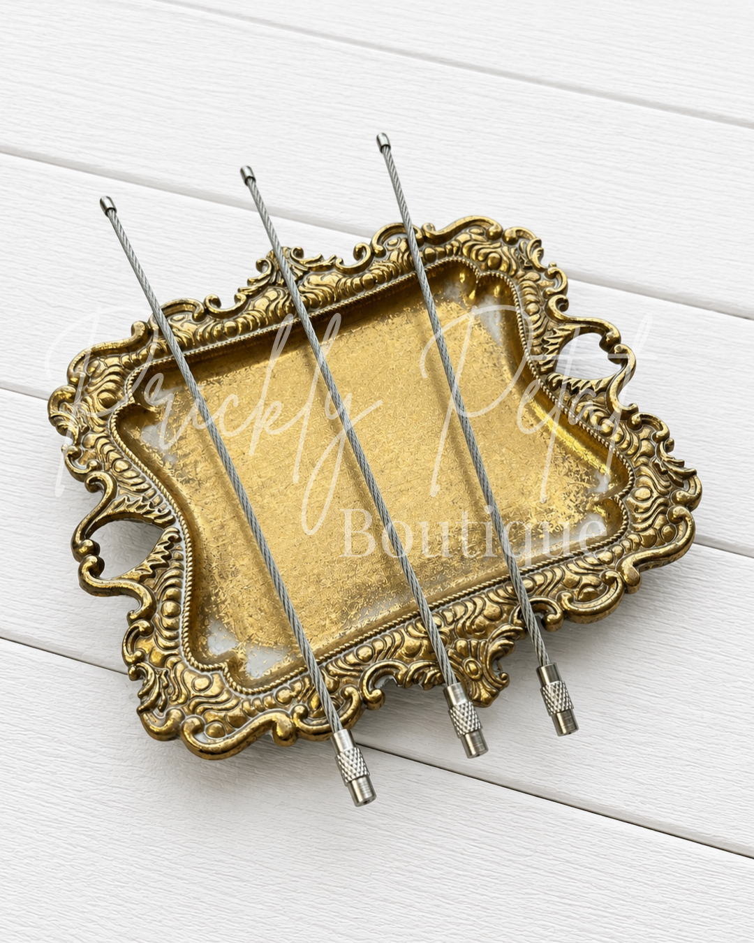 Four silver metal beadable car charm hooks with textured tips, arranged on an ornate gold-colored tray over a white wood plank background.
