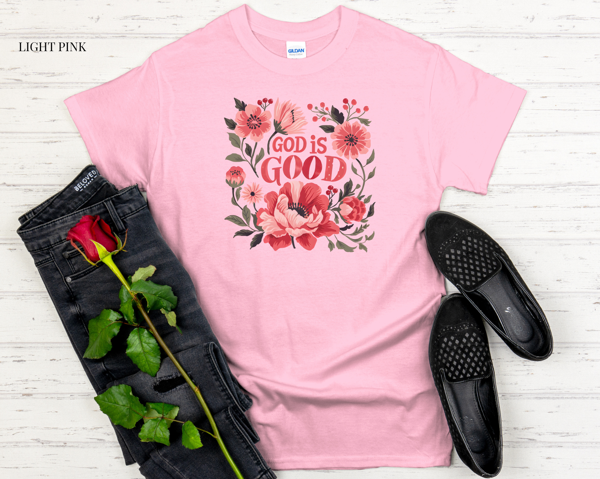 Light pink t-shirt with floral design and 'God is Good' text, paired with black jeans and shoes on a light wooden surface.