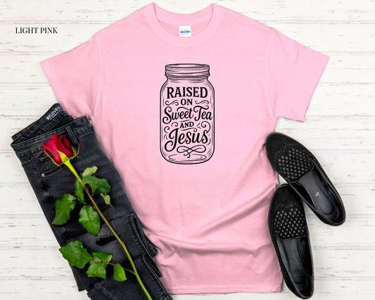Pink t-shirt with 'Raised on Sweet Tea and Jesus' design, paired with black jeans and shoes on a light wooden surface.