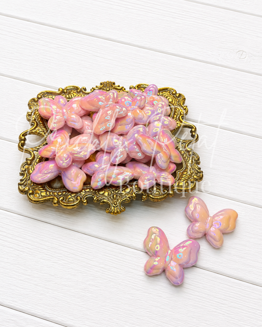 Pink iridescent butterfly-shaped beads displayed on an ornate gold tray, with two loose beads placed on a white wooden surface.
