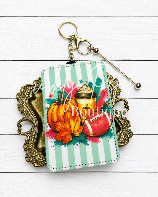 A rectangular wallet or card holder keychain with a mint green and white striped background, featuring a colorful autumn watercolor design with pumpkins, a football, and leaves. It is displayed on an ornate gold-colored tray over a white wood plank background.