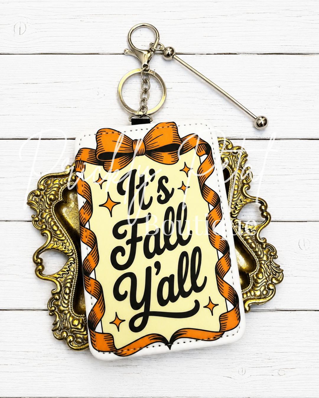 A square keychain or bag charm with a retro autumn design, featuring the text 'It's Fall Y'all' in black script on a cream background, surrounded by orange ribbons and stars. The charm is attached to a silver metal keyring and is displayed against a white wood plank background with an ornate gold frame behind it.