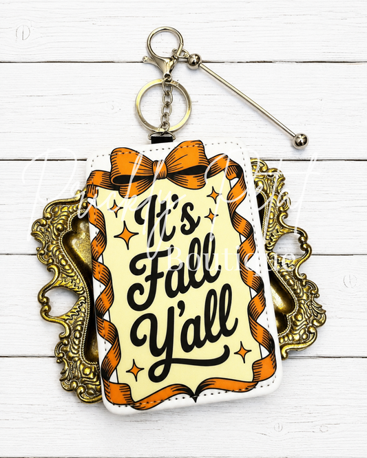 A square keychain or bag charm with a retro autumn design, featuring the text 'It's Fall Y'all' in black script on a cream background, surrounded by orange ribbons and stars. The charm is attached to a silver metal keyring and is displayed against a white wood plank background with an ornate gold frame behind it.