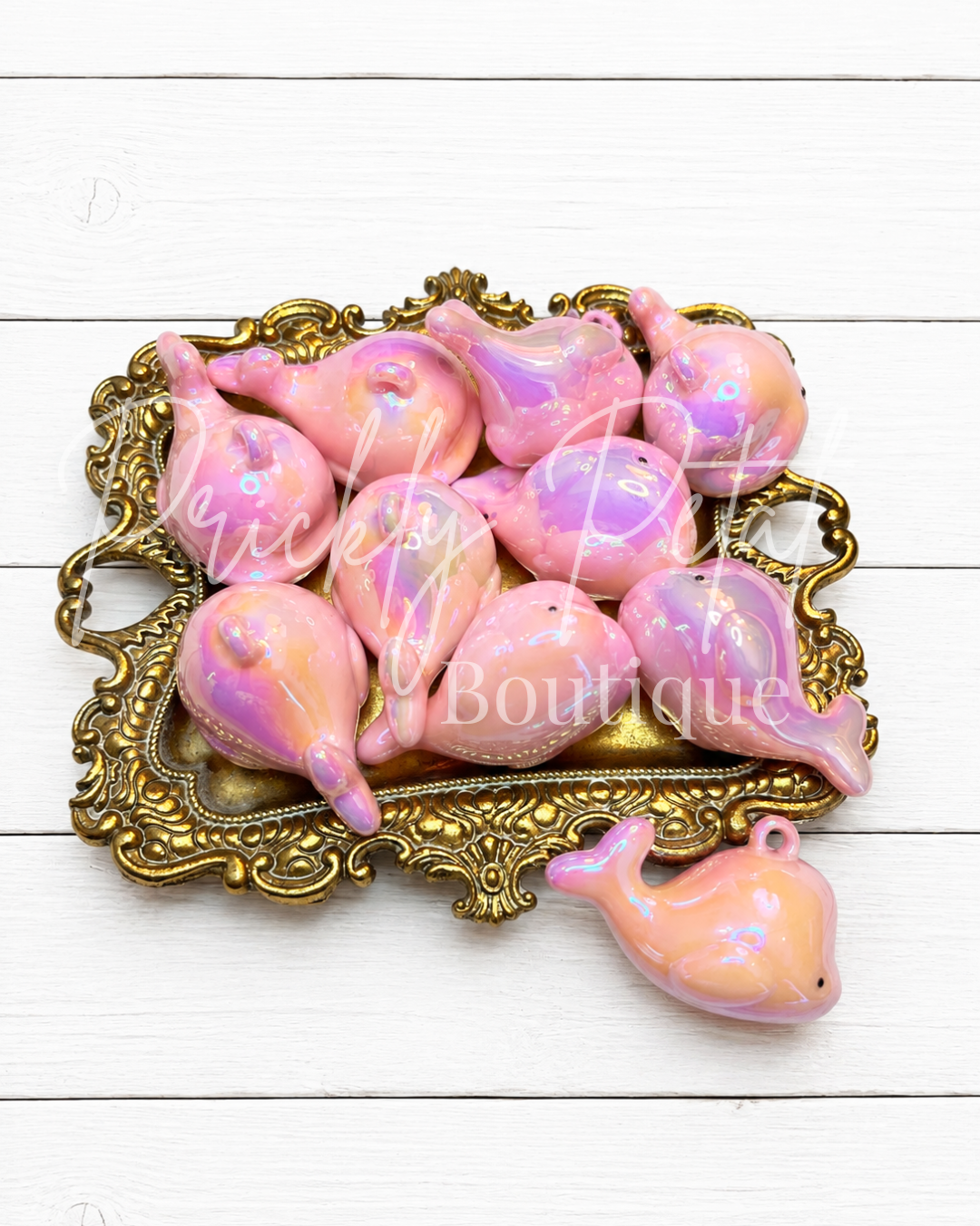 A set of glossy pink and purple seashell-shaped charms with an iridescent finish, displayed on an ornate gold tray with one charm placed in front on a white wood background.