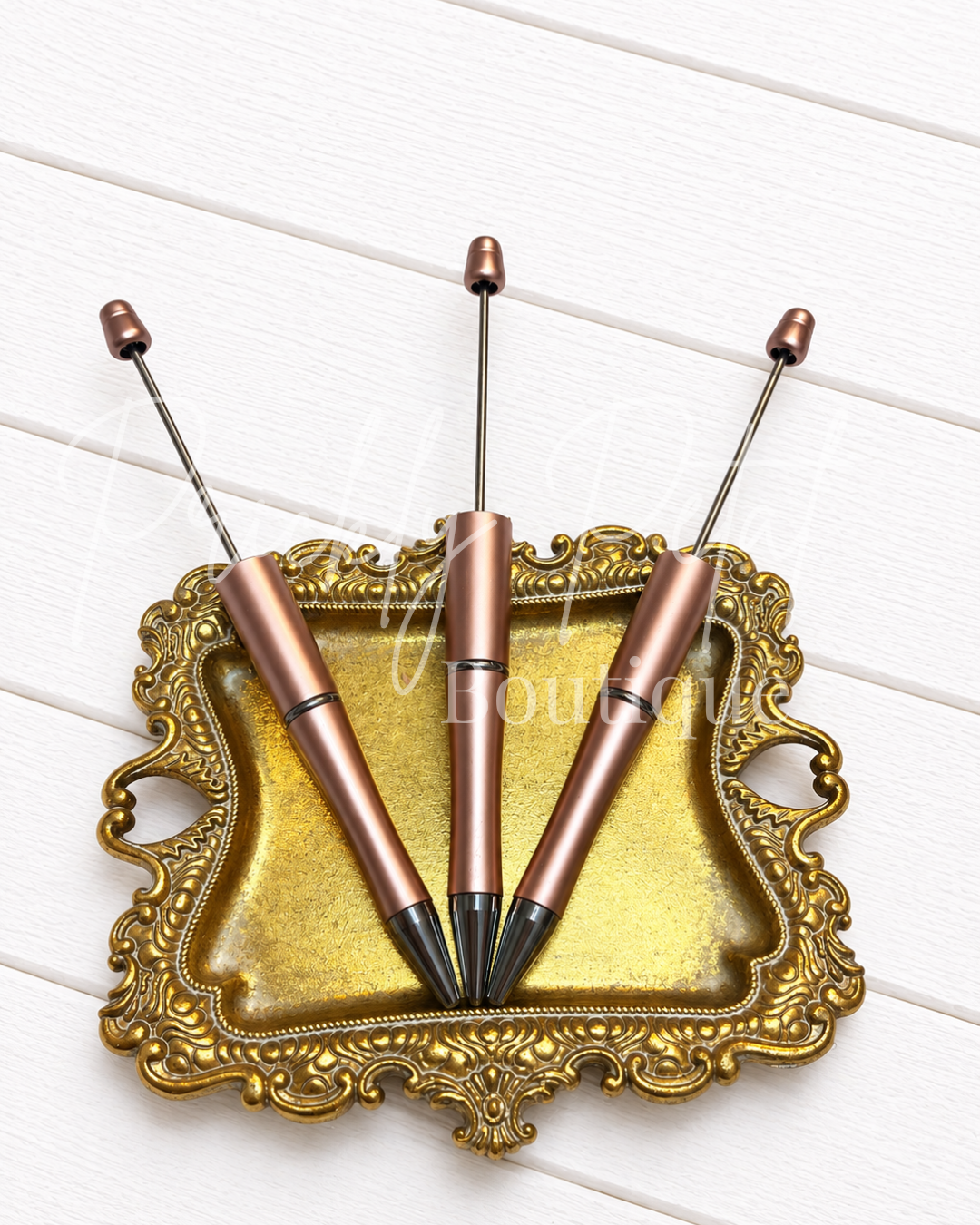 Three metallic rose gold pen-shaped tools with long silver rods and black tips, arranged on an ornate gold tray over a white wood plank background.