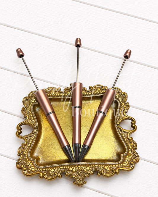 Three metallic rose gold pen-shaped tools with long silver rods and black tips, arranged on an ornate gold tray over a white wood plank background.