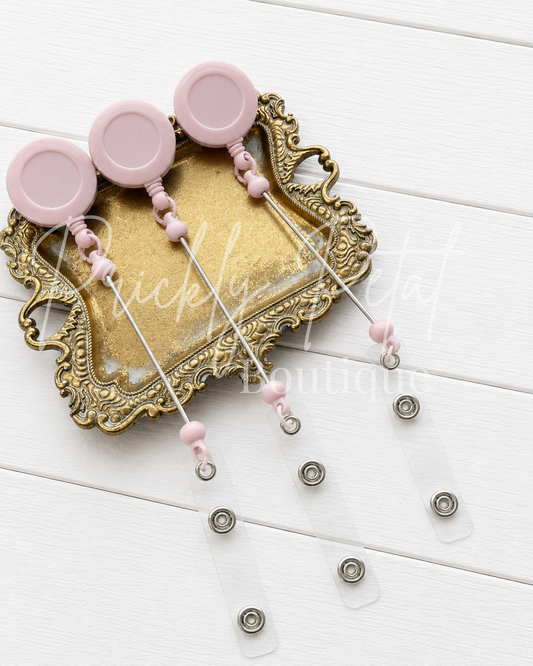 Three pink retractable badge reel cords with clear plastic ID tags and silver snap buttons, arranged on an ornate gold tray over a white wood background.
