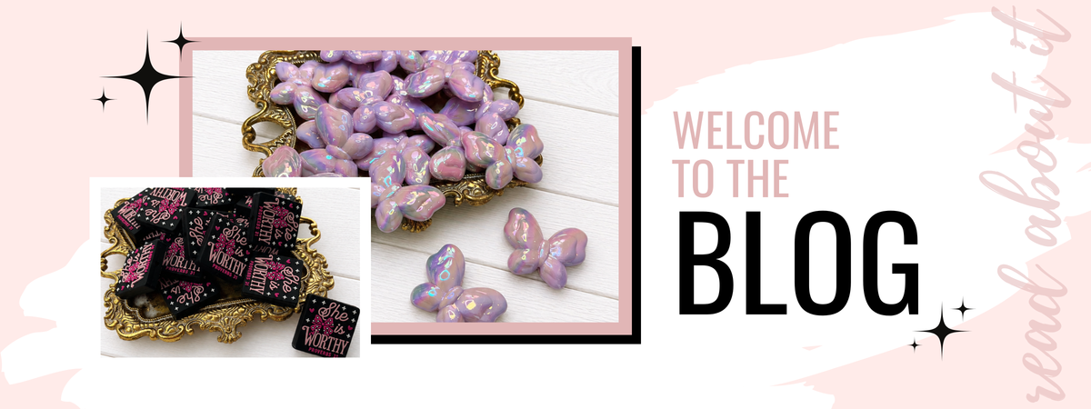 A promotional graphic for a blog featuring two inset photos of decorative pink and purple heart-shaped beads or charms displayed in gold trays, alongside the text 'WELCOME TO THE BLOG' on a pale pink background.