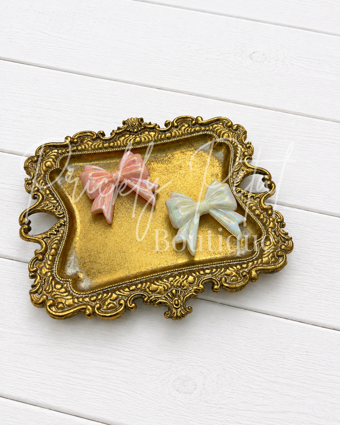 An ornate gold-colored decorative tray with two small bow charms, one pink and one pale green, displayed on a white wooden background.