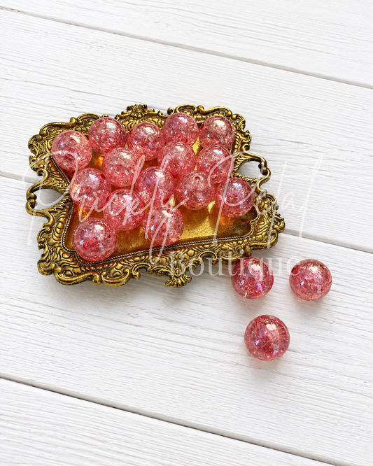 Pink glittery round beads displayed in an ornate gold-colored tray on a white wooden background, with three additional beads placed beside the tray.