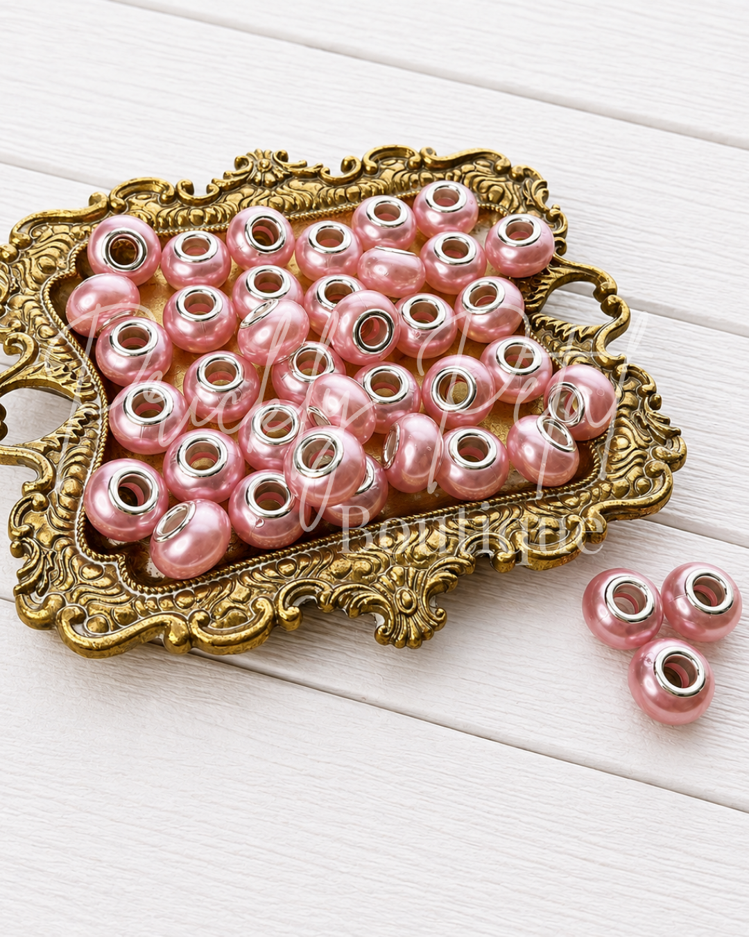 Pink faux pearl spacer beads with silver metal ends, displayed on an ornate gold tray over a white wood background.