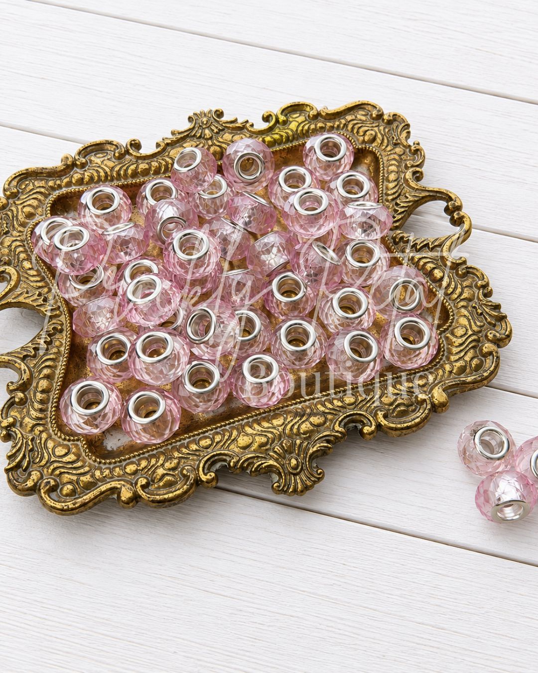 Pink translucent glass beads with silver metal spacers, displayed on an ornate gold tray over a white wooden background.