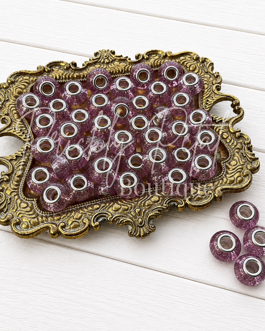 A decorative tray filled with round purple beads with silver metal rims, alongside four loose matching beads on a white wood background.