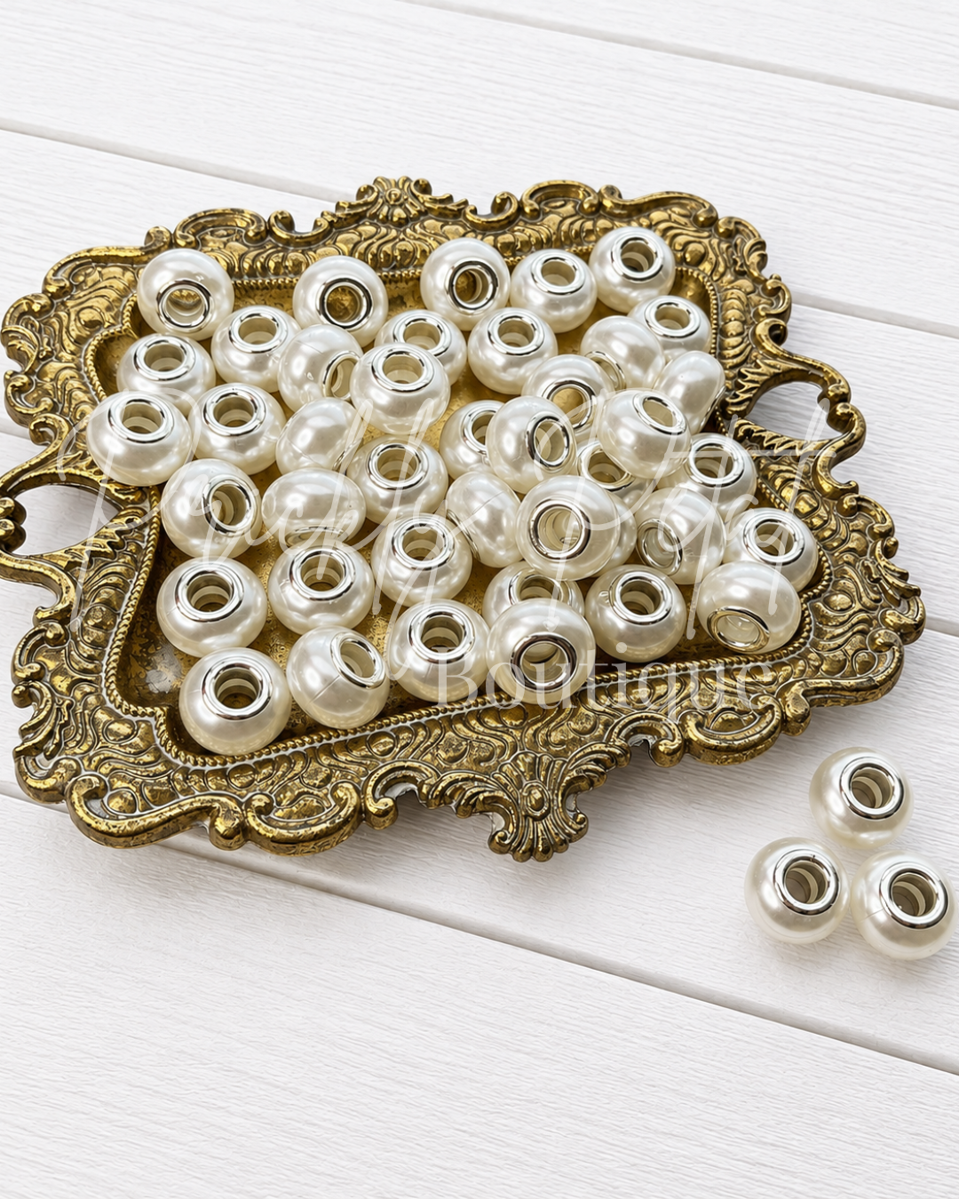 A pile of small round pearl-like beads with silver-toned metal ends, displayed on an ornate gold tray on a white wood surface.