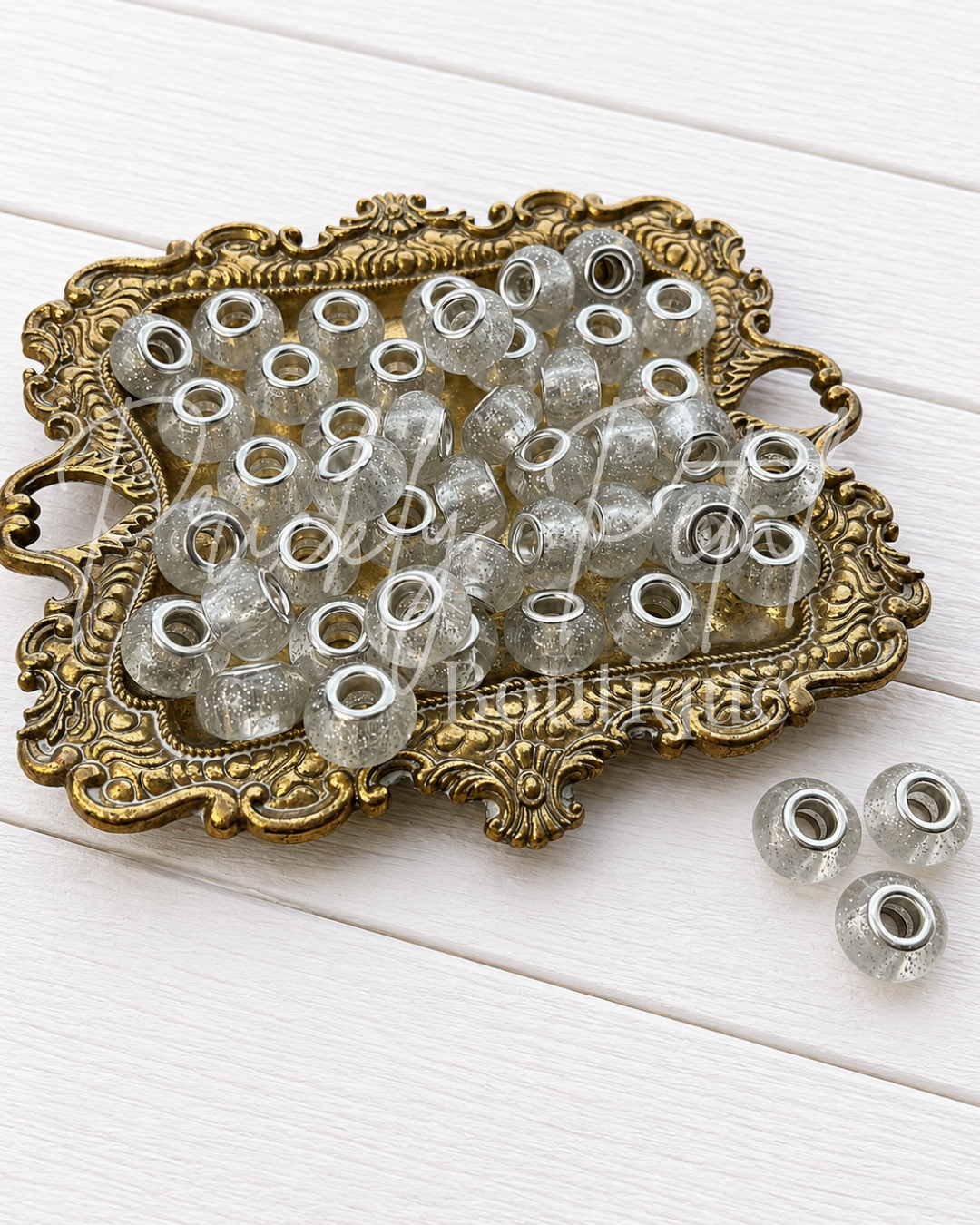 Clear glass seed beads with silver glitter and silver metal eyelets, displayed on an ornate gold tray with three additional beads on a white wooden background.