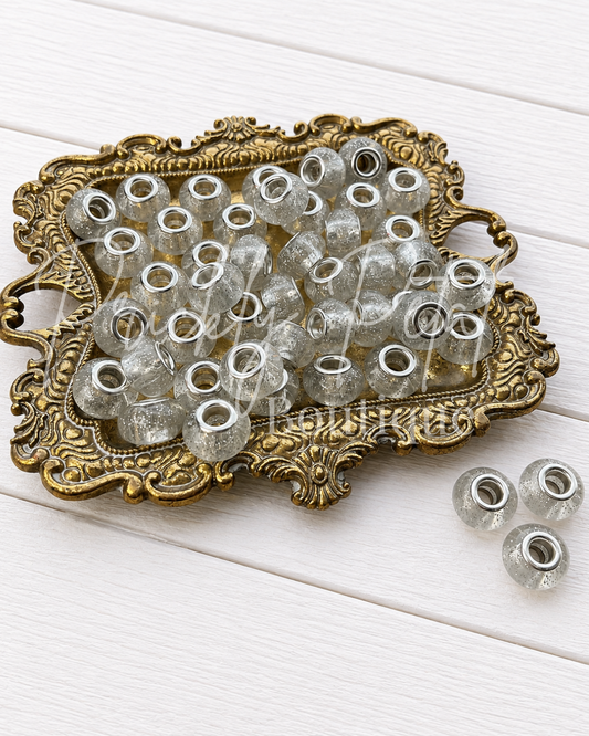 Clear glass seed beads with silver glitter and silver metal eyelets, displayed on an ornate gold tray with three additional beads on a white wooden background.