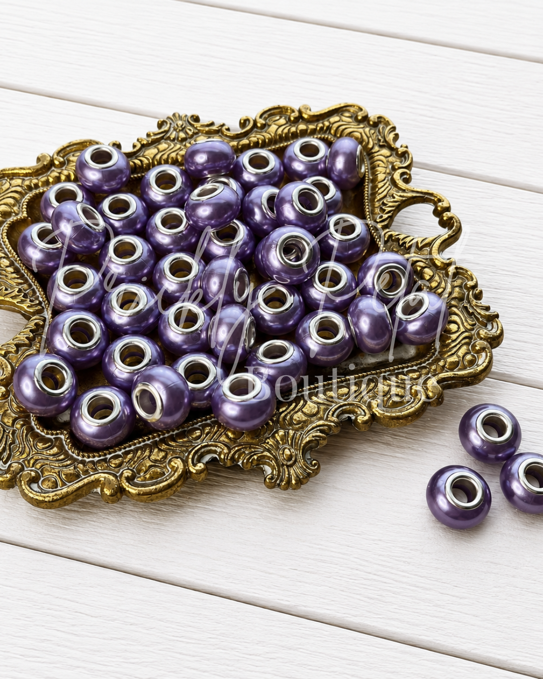Purple metallic round beads with silver-toned metal cores, displayed on an ornate gold tray over a white wood plank background.