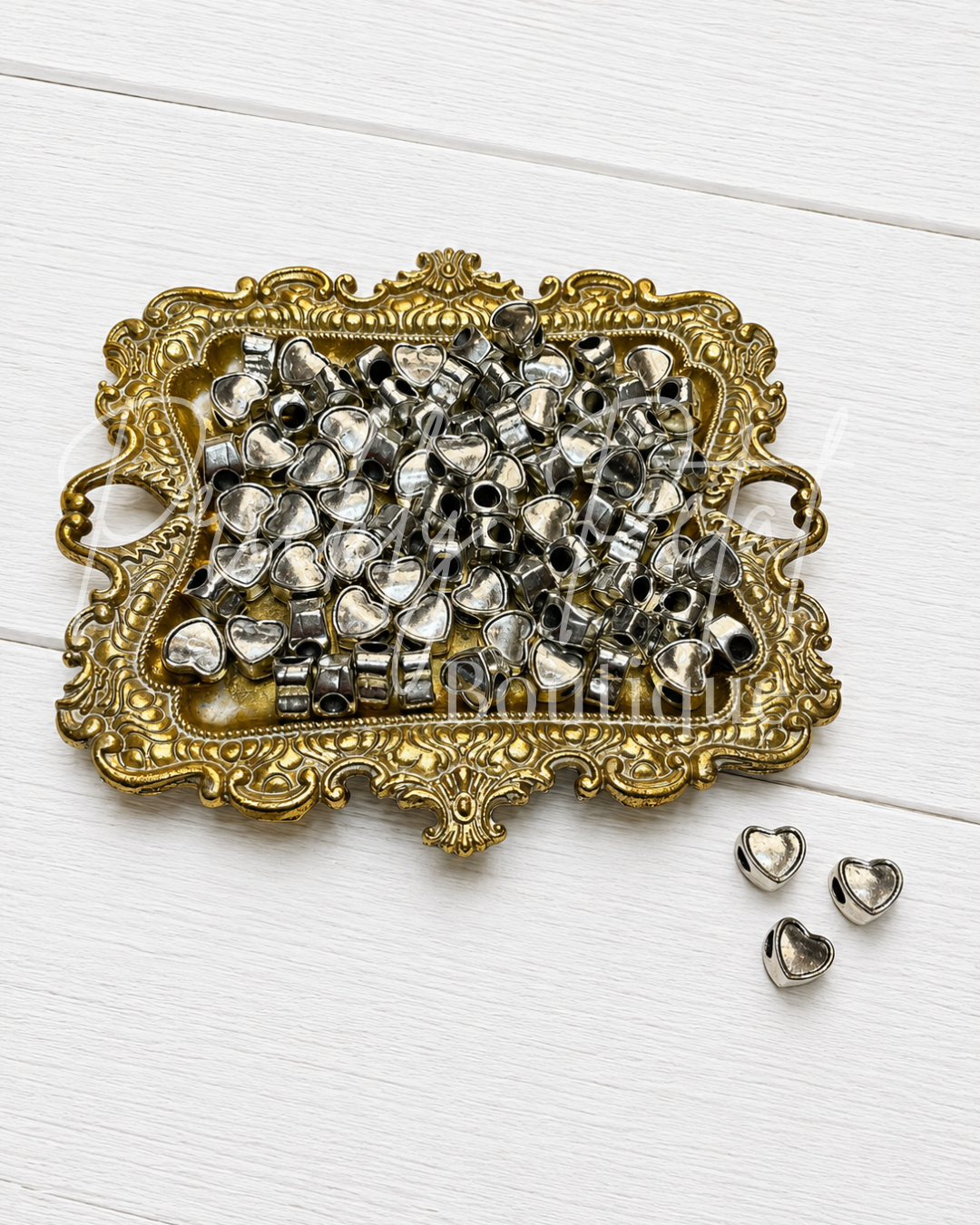 A pile of small silver heart-shaped beads with metal loops, displayed on an ornate gold tray with three individual beads placed on a white wooden surface.