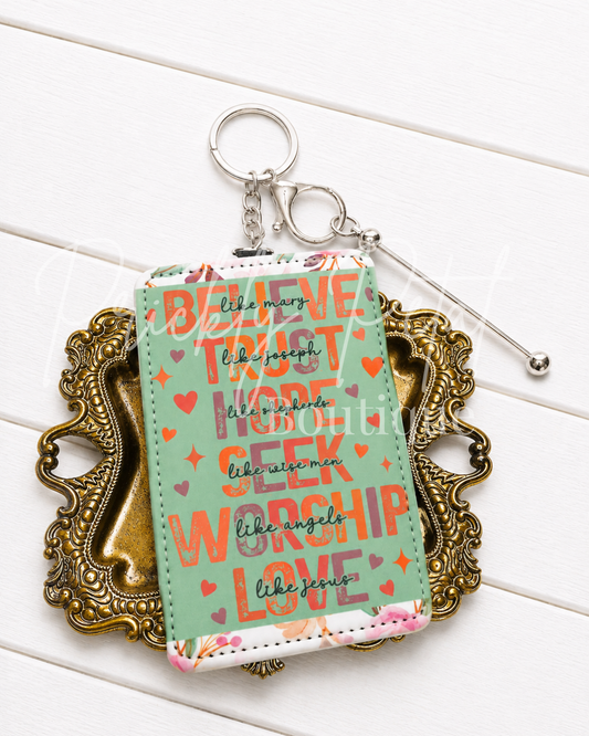 A mint green keychain wallet with a floral border and inspirational religious text, displayed on an ornate gold-colored frame over a white wood background.