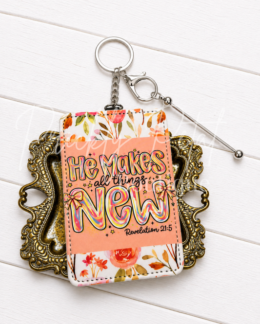 A floral and text keychain pouch with the phrase 'He makes all things new - Revelation 21:5' in colorful lettering, displayed on an ornate gold tray over a white wood background.