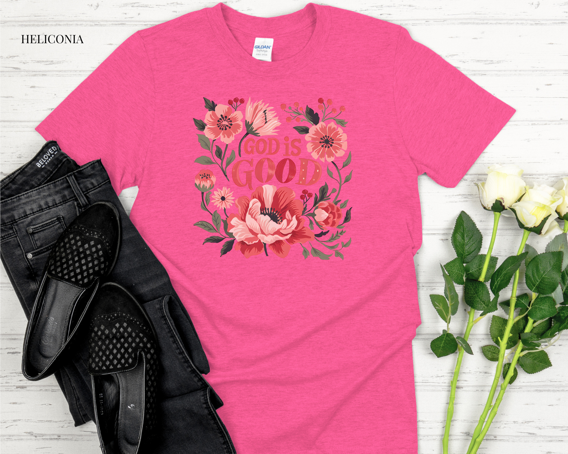 Pink t-shirt with floral design and 'God is Good' text on a white wooden surface with black shoes and flowers.