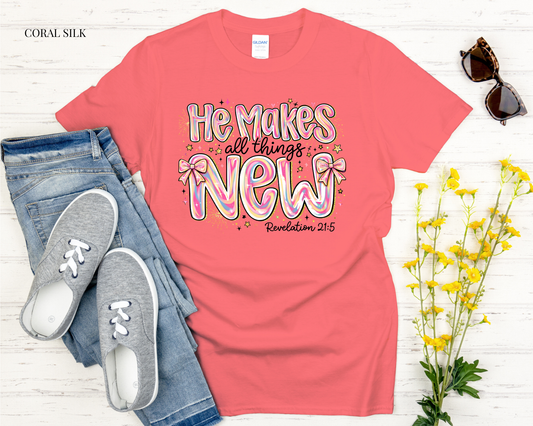 Coral t-shirt with 'He makes all things new' text, paired with jeans and sneakers on a light wooden surface.
