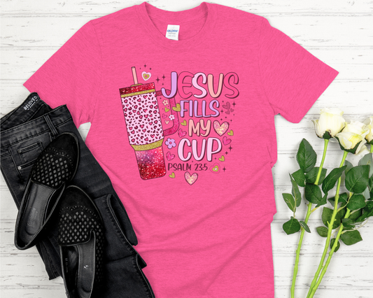 Pink t-shirt with 'Jesus Fills My Cup' design on a white wooden surface with black shoes and flowers.
