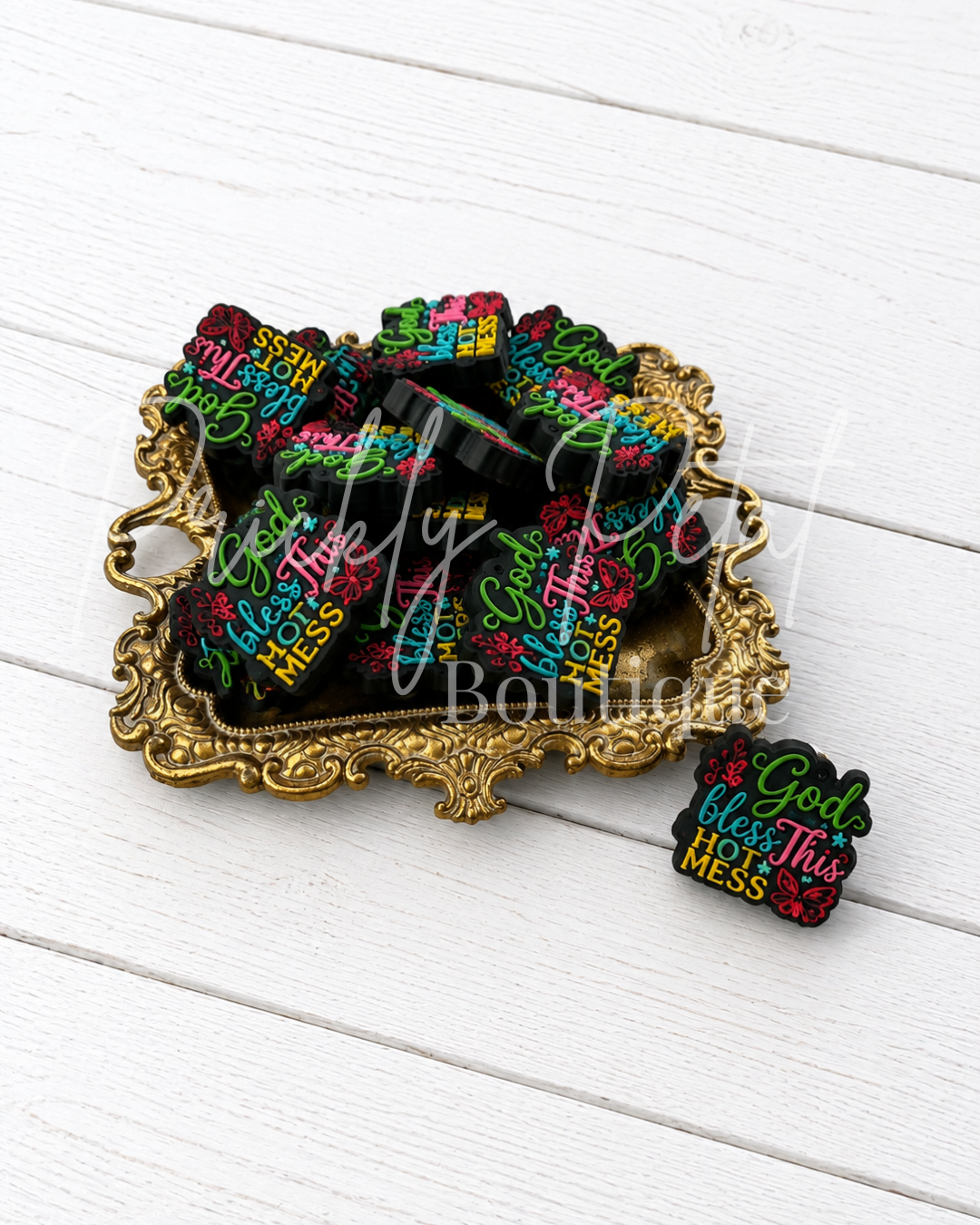 silicone focal beads with colorful text reading 'God bless this mess' in pink, green, yellow, and white, displayed on an ornate gold tray with one bead placed beside it on a white wood plank background.