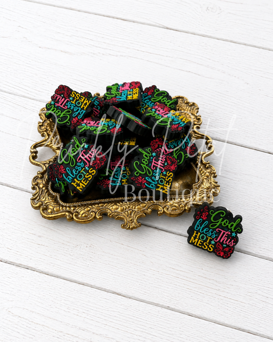 silicone focal beads with colorful text reading 'God bless this mess' in pink, green, yellow, and white, displayed on an ornate gold tray with one bead placed beside it on a white wood plank background.