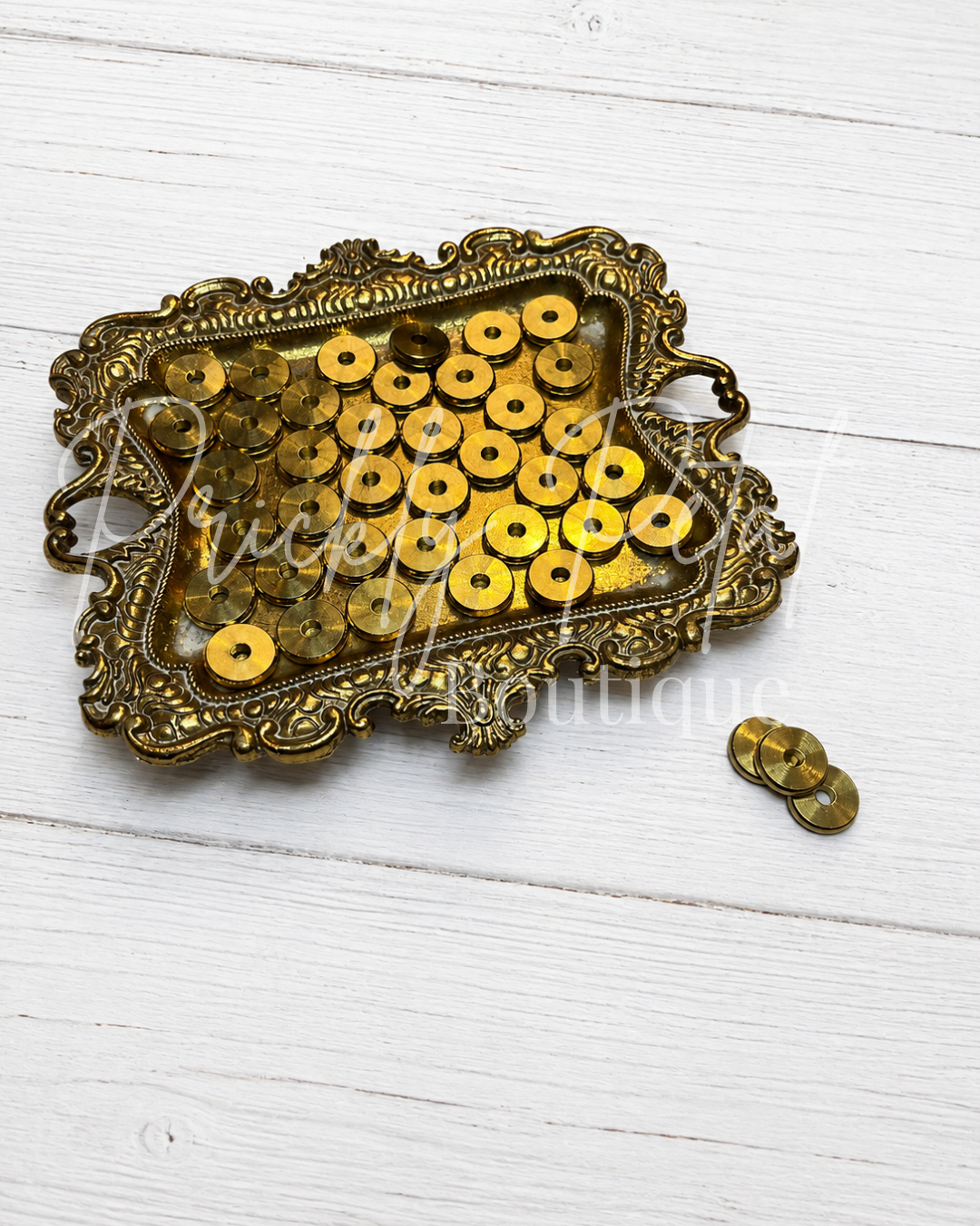 A vintage-style ornate gold tray filled with round gold spacer bead, with a few loose spacer bead beside it, photographed on a white wood plank background.