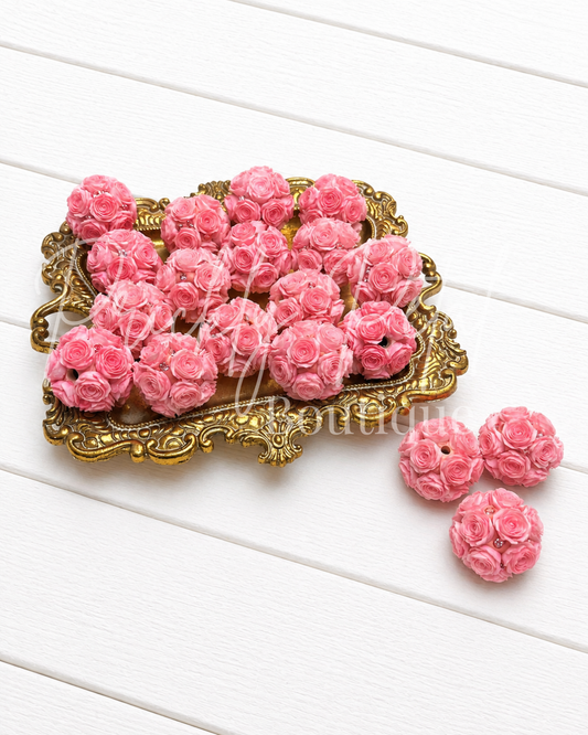 Pink clay beads shaped like roses, displayed on an ornate gold tray with three additional beads placed beside it on a white wood plank background.