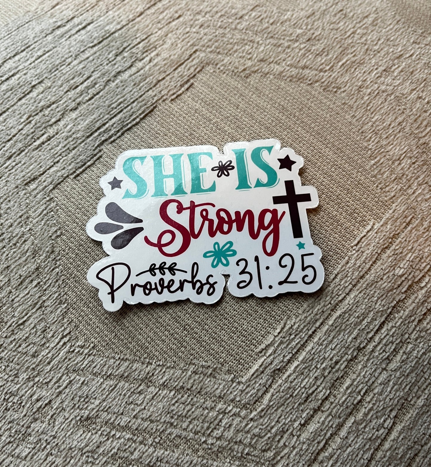 Sticker with 'SHE IS Strong Proverbs 31:25' text on a textured surface