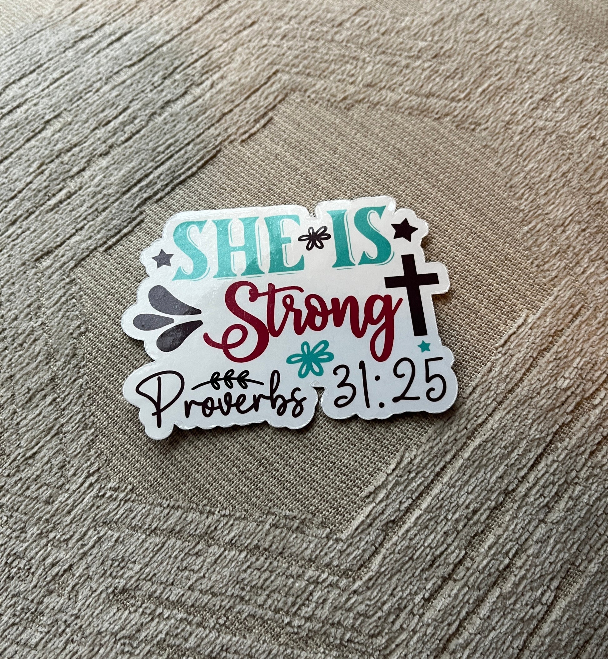Sticker with 'SHE IS Strong Proverbs 31:25' text on a textured surface